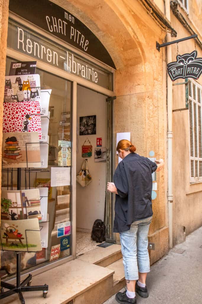 A black cat sits in the doorway at the entrance of Mon Chat Pitre while a a customer enters the store.