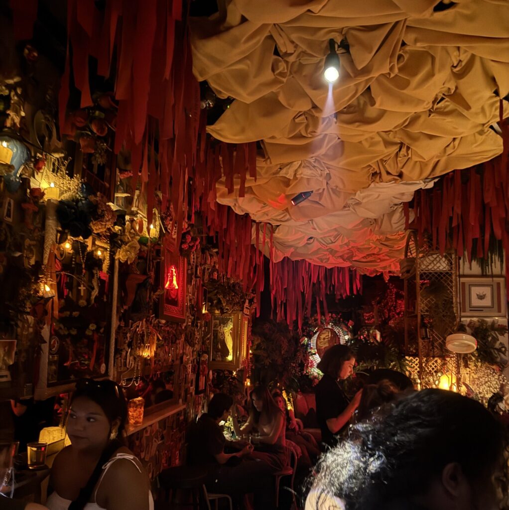 Image of the inside of a bar that is softly lit with fabric adorning the ceiling and people mingling all around.
